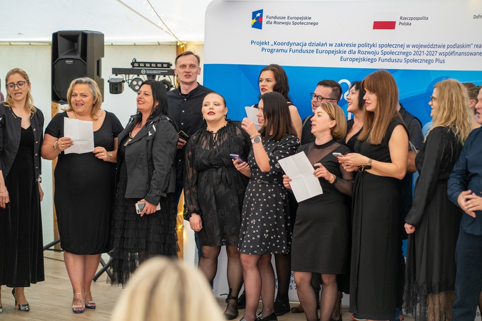 Zdjęcie przedstawia osoby stojące na dworze w tym kobiety i mężczyzn, którzy śpiewają przed publicznością