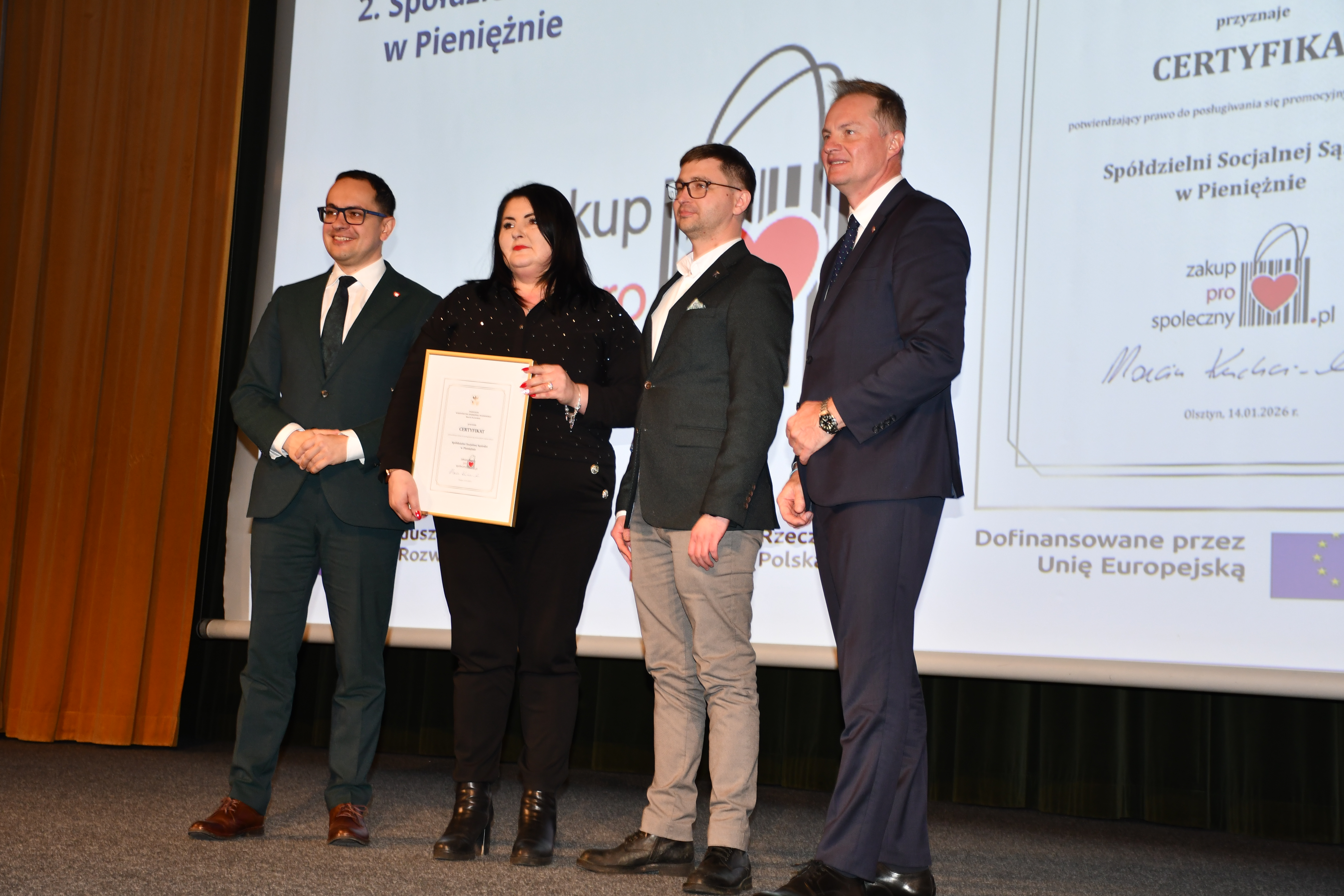 Zdjęcie przedstawia osoby stojące na scenie zarówno mężczyźni jak i kobiety, które trzymają w ręku ramki z certyfikatami Znaku Zakup Prospołeczny
