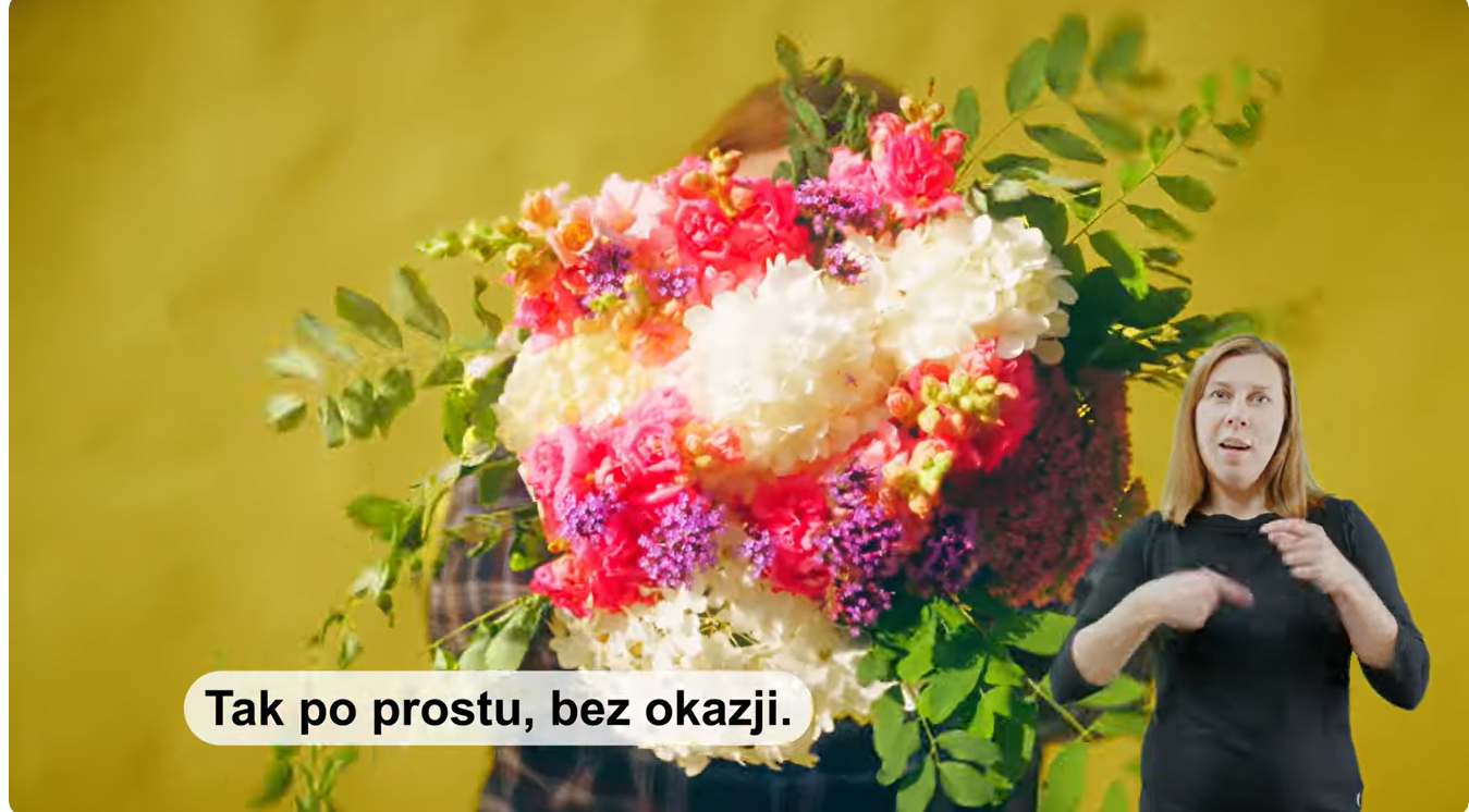 Zdjęcie przedstawia kolorowy bukiet kwiatów, poniżej z prawej strony kobieta tłumaczy na Polski Język Migowy. Na samym dole widnieje napis z klatki filmu 