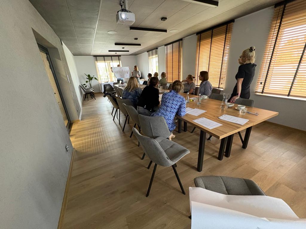 Zdjęcie przedstawia sale szkoleniową, w której znajduję się długi stół przy którym siedzą osoby zarówno kobiety jak i mężczyźni