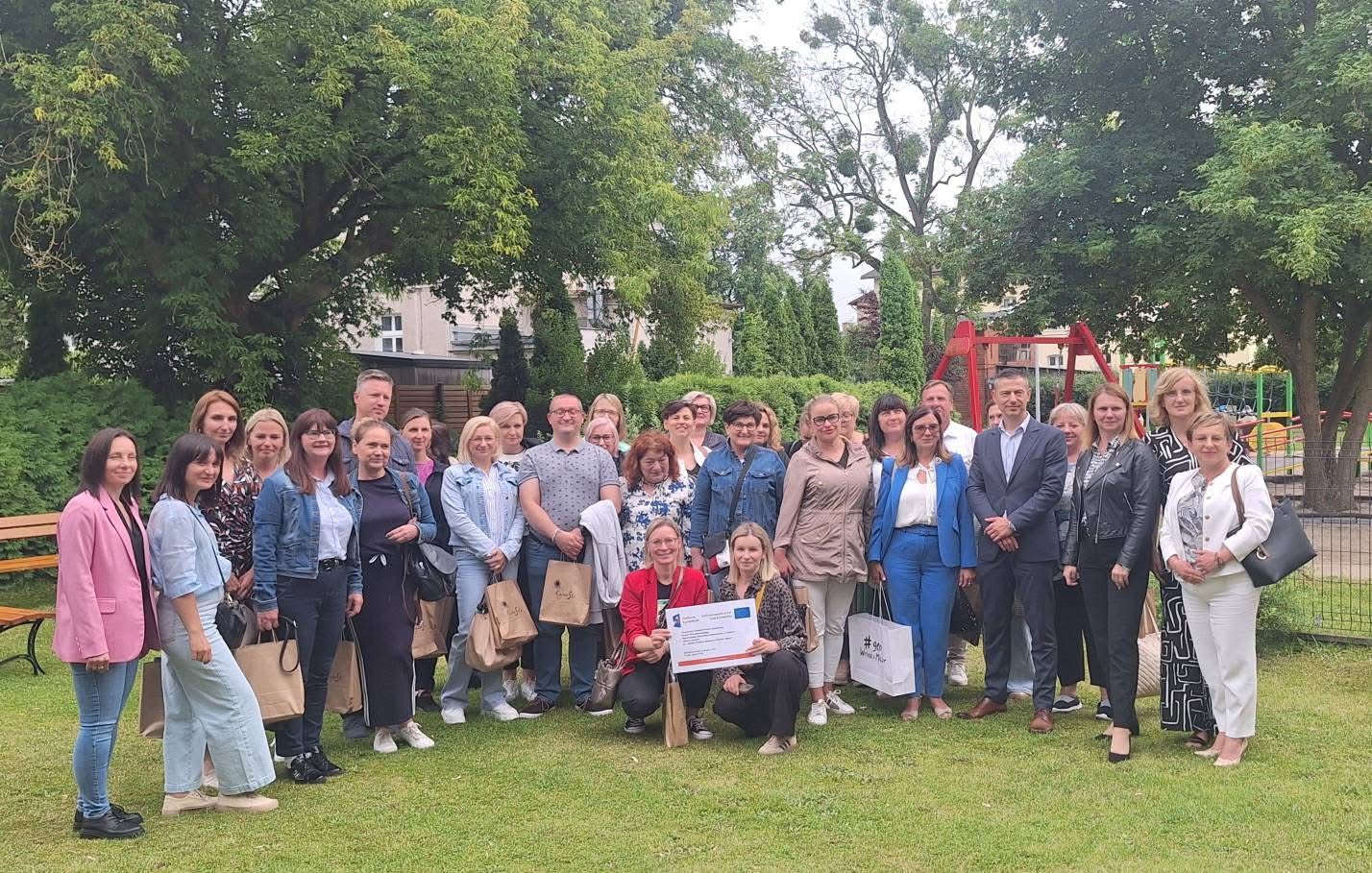 Zdjęcie przedstawia osoby stojące w parku na zielonej trawie wokół pełno drzew. Wśród tych osób są zarówno kobiety jak i mężczyźni.