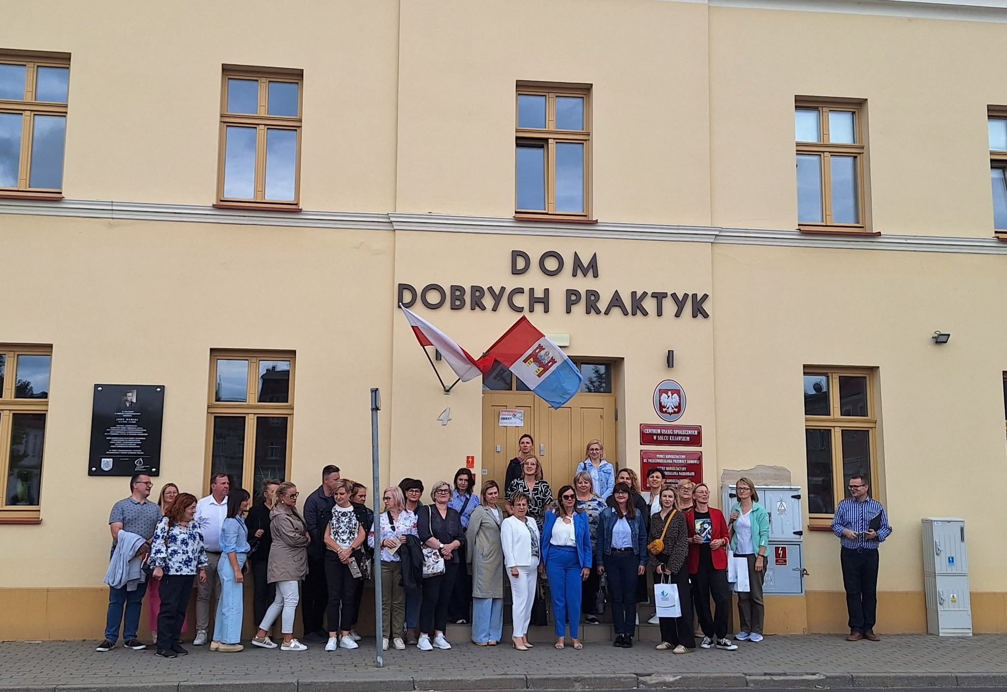 Zdjęcie przedstawia osoby stojące przed budynkiem wśród których są zarówno kobiety jak i mężczyzn