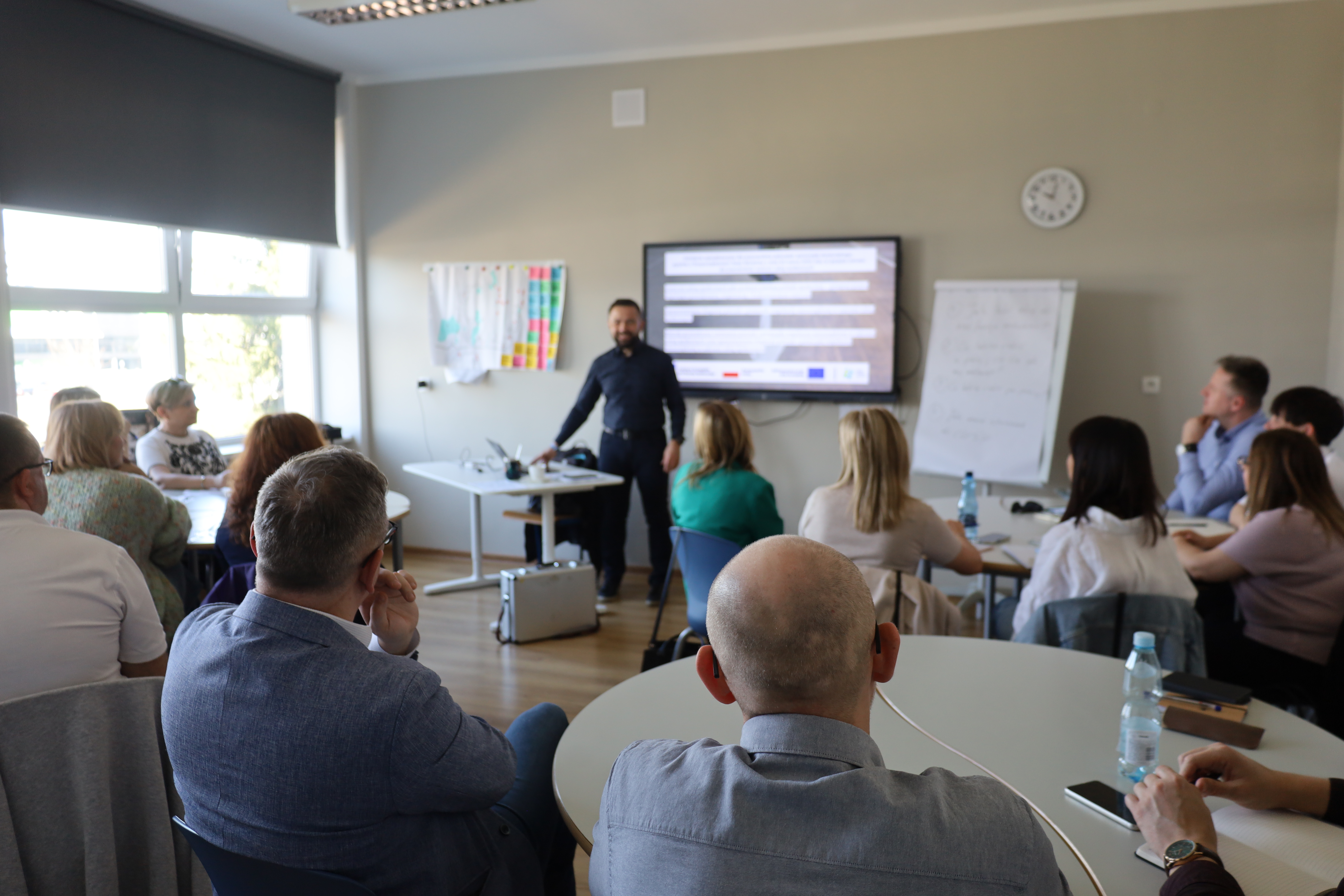 Zdjęcie przedstawia uczestników szkolenia siedzących przy okragłym stole i obserwujących prowadzącego zajęcia. W tle widać ekran z prezentacją.