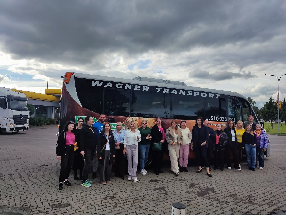 Zdjęcie przedstawia osoby wśród nich są zarówno kobiety i mężczyźni stojące przed autobusem na jakimś parkingu
