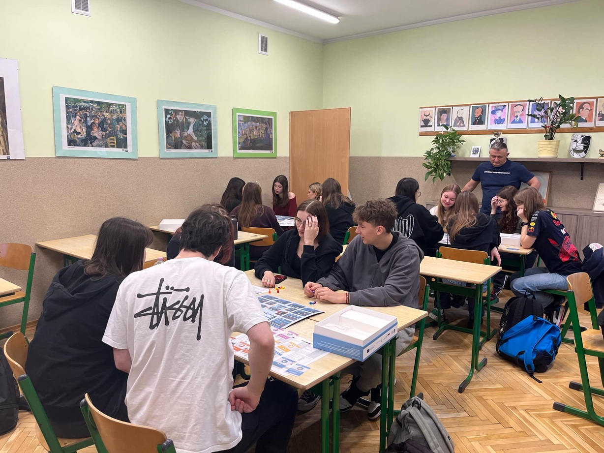 Zdjęcie przedstawia młode osoby uczniów szkoły, którzy siedzą w ławce szkolnej i grają w planszową  dot. ekonomii społecznej